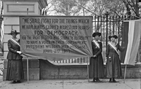 A black and white photograph of three suffragists standing in front of the White House holding a banner that says: "We shall fight for things which we have always carried nearest our hearts—for democracy, for the right of those who submit to authority to have a voice in their government."
